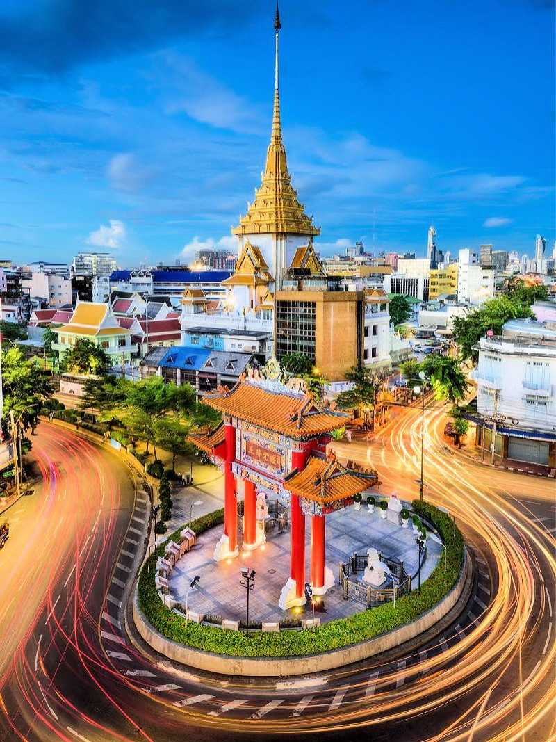 Goldener Tempel und Tor im historischen Stadtteil von Bangkok, Thailand, mit Lichtspuren von vorbeifahrenden Autos bei Sonnenuntergang