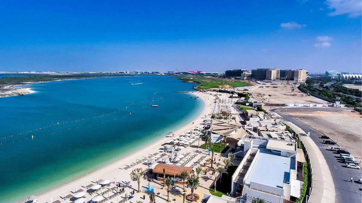 Luftaufnahme eines luxuriösen Strandes mit Liegestühlen, türkisfarbenem Wasser und modernen Gebäuden im Hintergrund, bei strahlend blauem Himmel